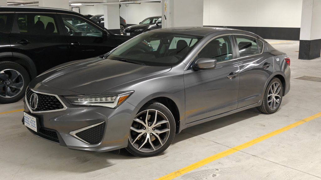 A grey Acura ILX stands handsomely in a covered parking lot with bright lights, with other cars in the background. The front wheels of the car are turned, as if it is posing for the photo with a smirk. The license plate reads EEEE EEE.