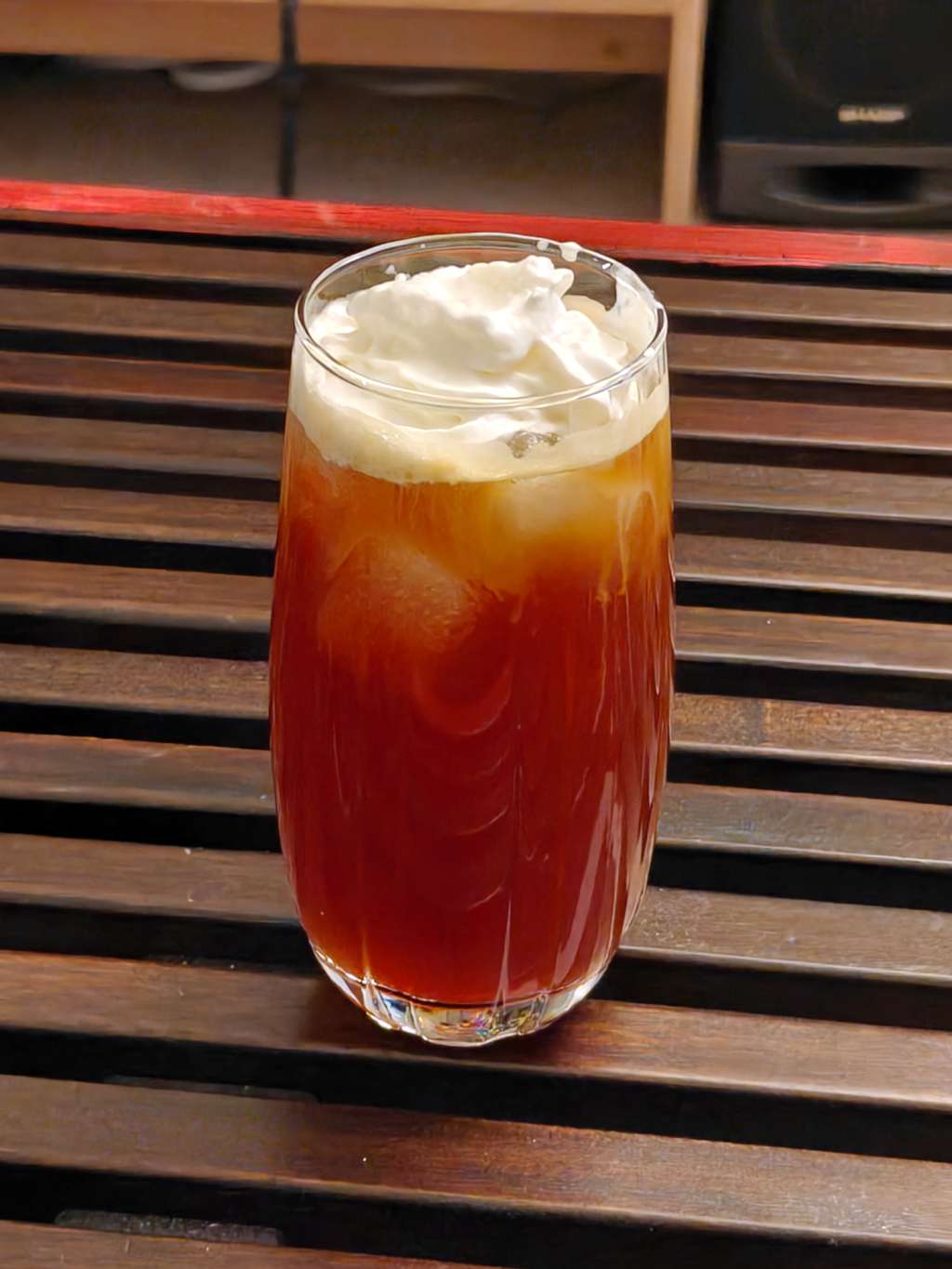 A light-brown drink with ice inside and whipped cream on top sits on a table made of wooden slats, in a tall glass.