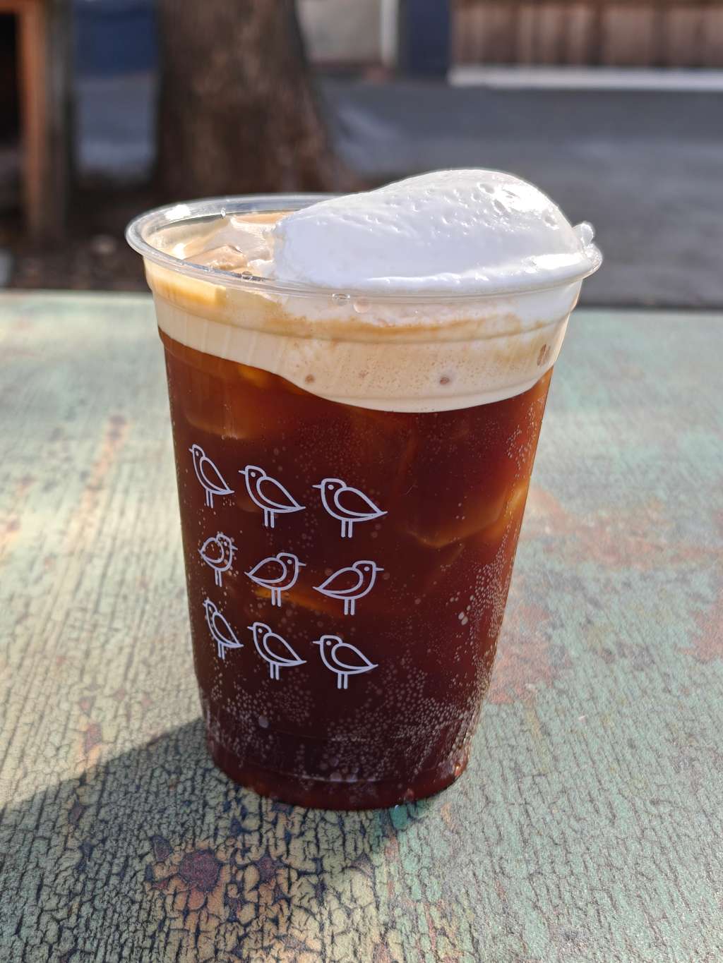 A dark brown drink with ice inside and whipped cream on top sits on a wooden table. The plastic cup has nine birds drawn on it, arranged in a grid shape.